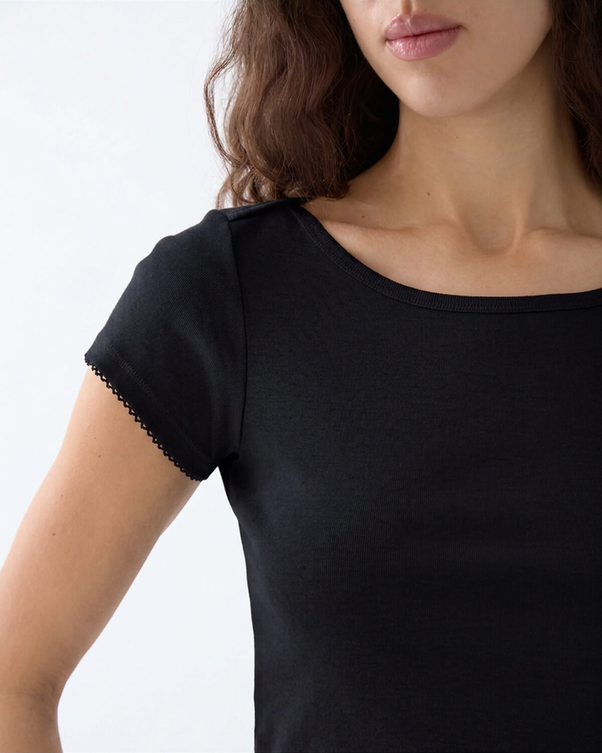 Close-up of a woman wearing a black short-sleeve top with a scoop neckline and delicate scalloped trim along the sleeve edge, against a plain light background.