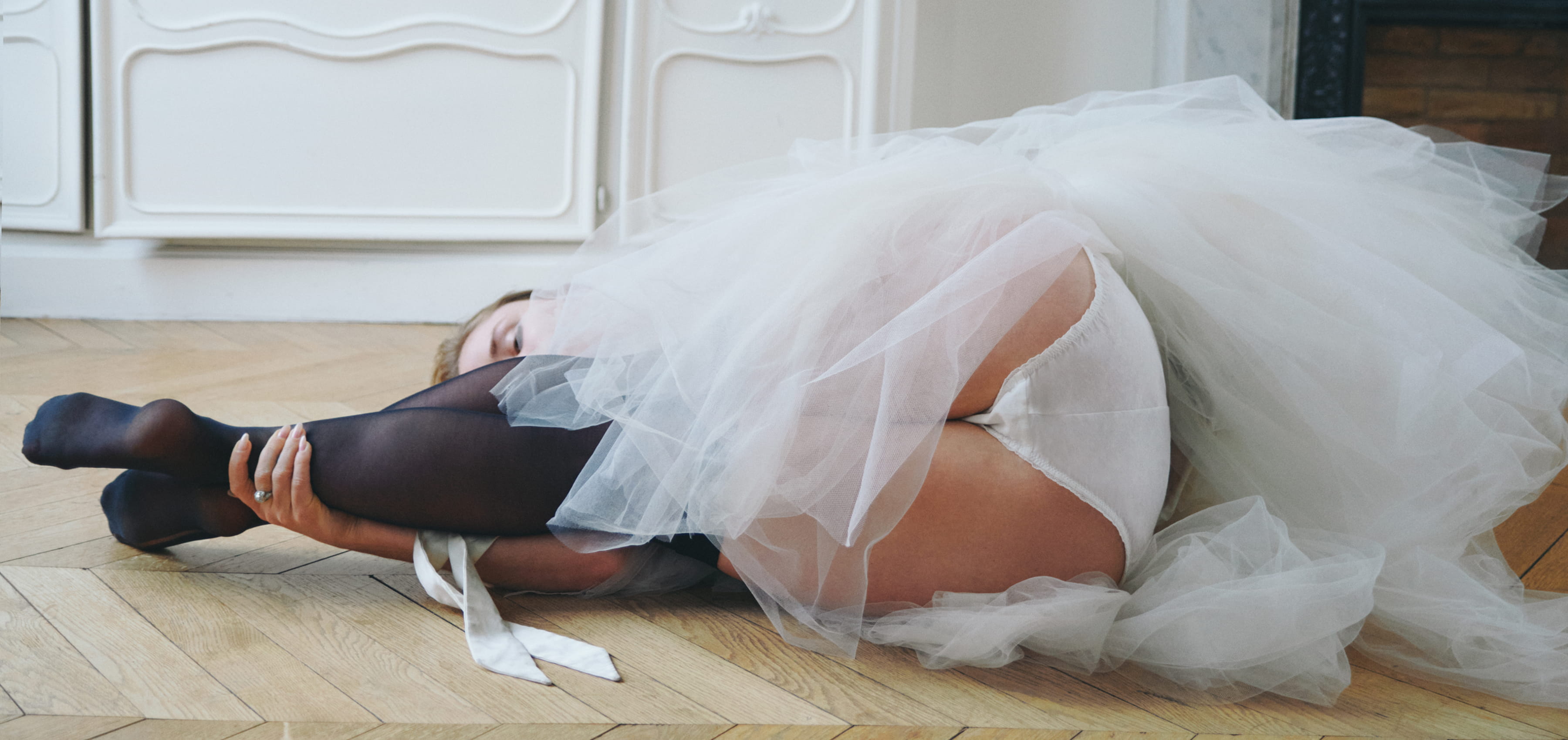 Woman lying on a wooden floor in a white tulle skirt and white underwear. 