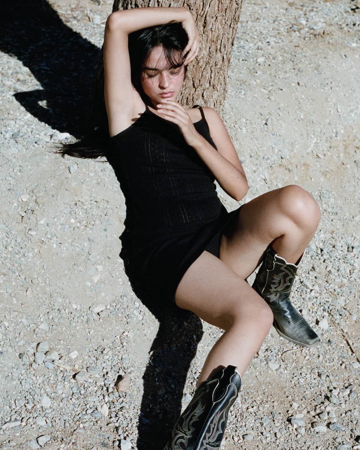 Woman in a black dress and boots leaning against a tree on a gravel surface