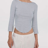 A woman wearing a cropped, three-quarter sleeve top with thin navy and white horizontal stripes, paired with white drawstring pants, against a plain light gray studio background.