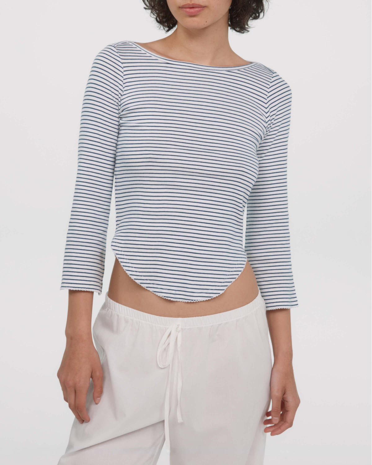 A woman wearing a cropped, three-quarter sleeve top with thin navy and white horizontal stripes, paired with white drawstring pants, against a plain light gray studio background.