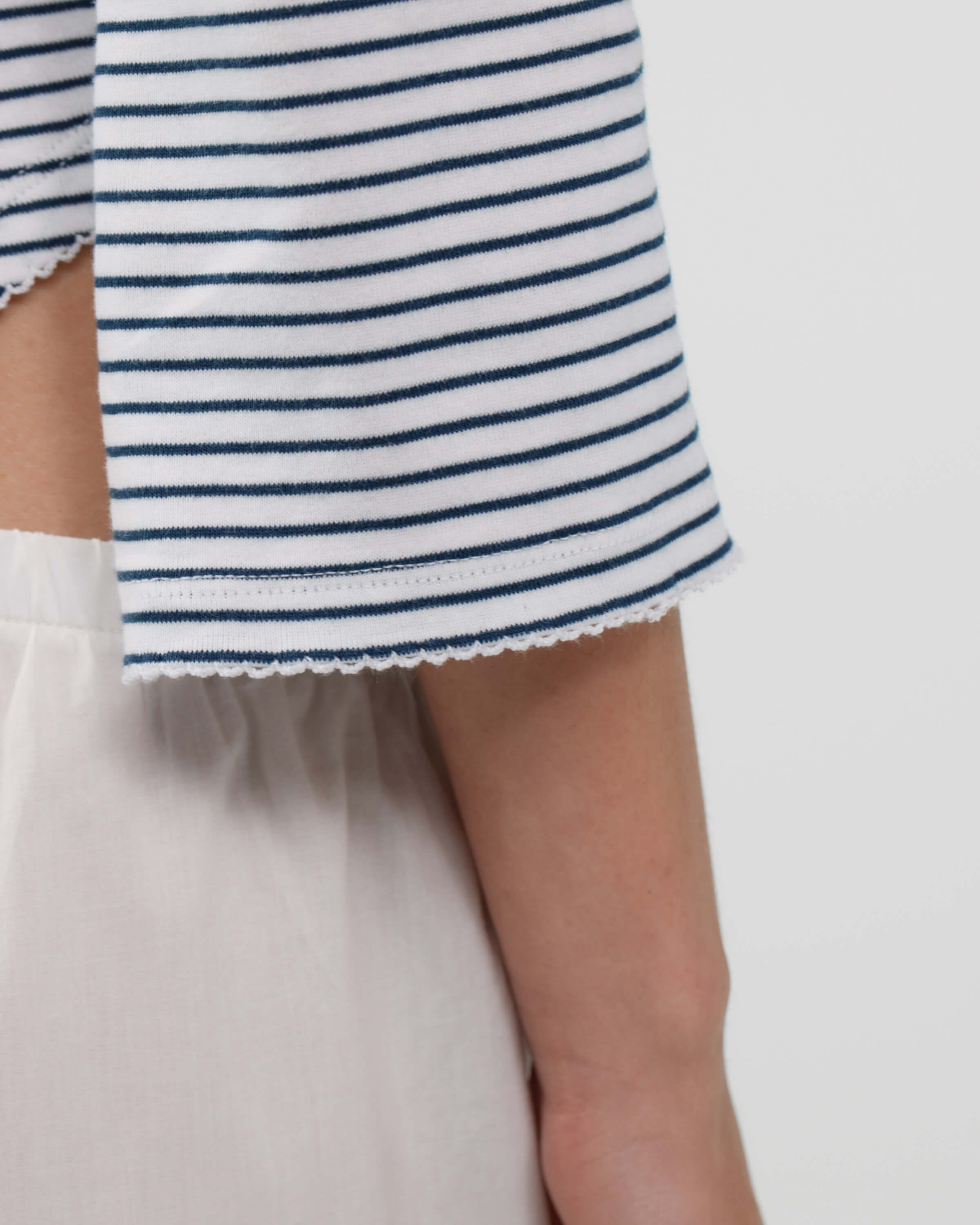 Close-up of a person wearing a white top with thin navy horizontal stripes and a scalloped hem, paired with white bottoms, against a plain light background.