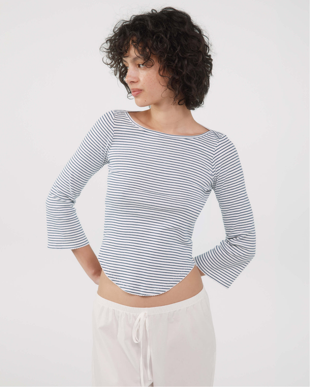 A woman with short curly dark hair wearing a blue and white horizontally striped long-sleeve crop top and white drawstring pants, photographed against a plain light gray studio background.
