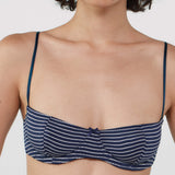 A close-up of a model wearing a navy blue and white striped bra with thin shoulder straps and a small front tie detail, set against a plain light gray background.