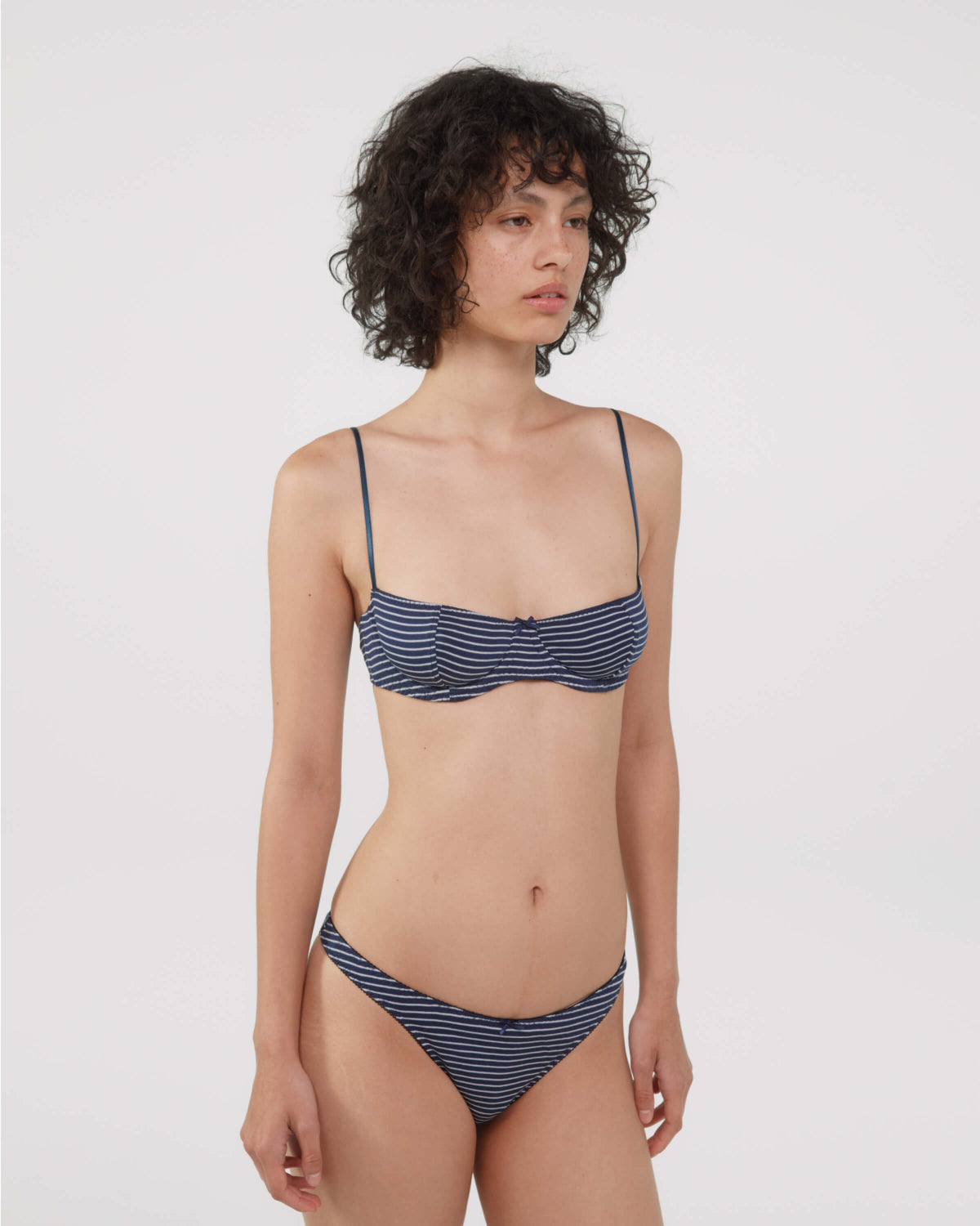 A model wearing a bra and thong with navy blue and white horizontal stripes, photographed against a solid light gray background.