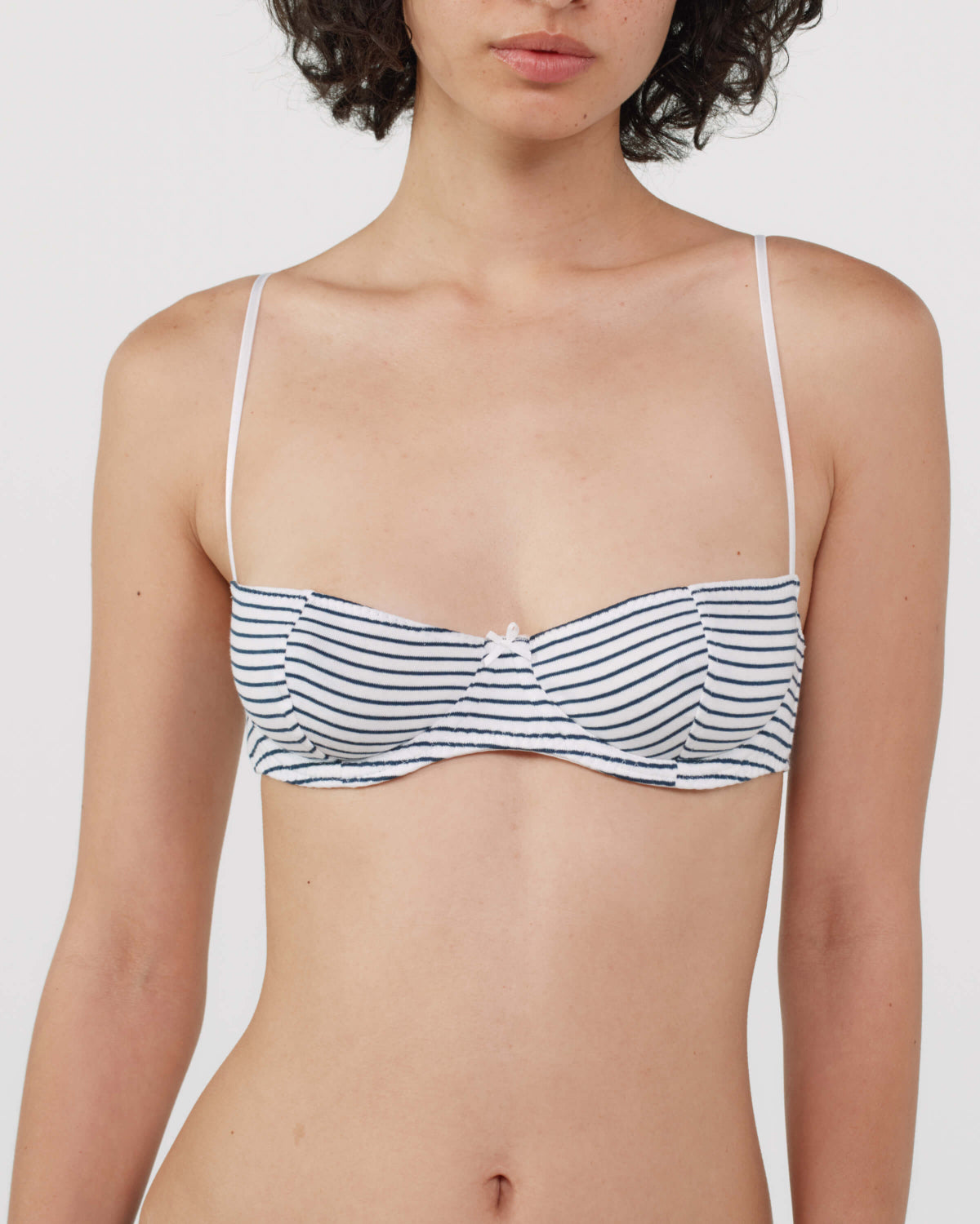 A close-up of a model wearing a white bra with thin shoulder straps and horizontal navy blue stripes, set against a plain light gray background.