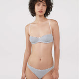 A model wearing a bra and thong with thin white straps and a blue and white horizontal stripe pattern, photographed against a solid light gray background.
