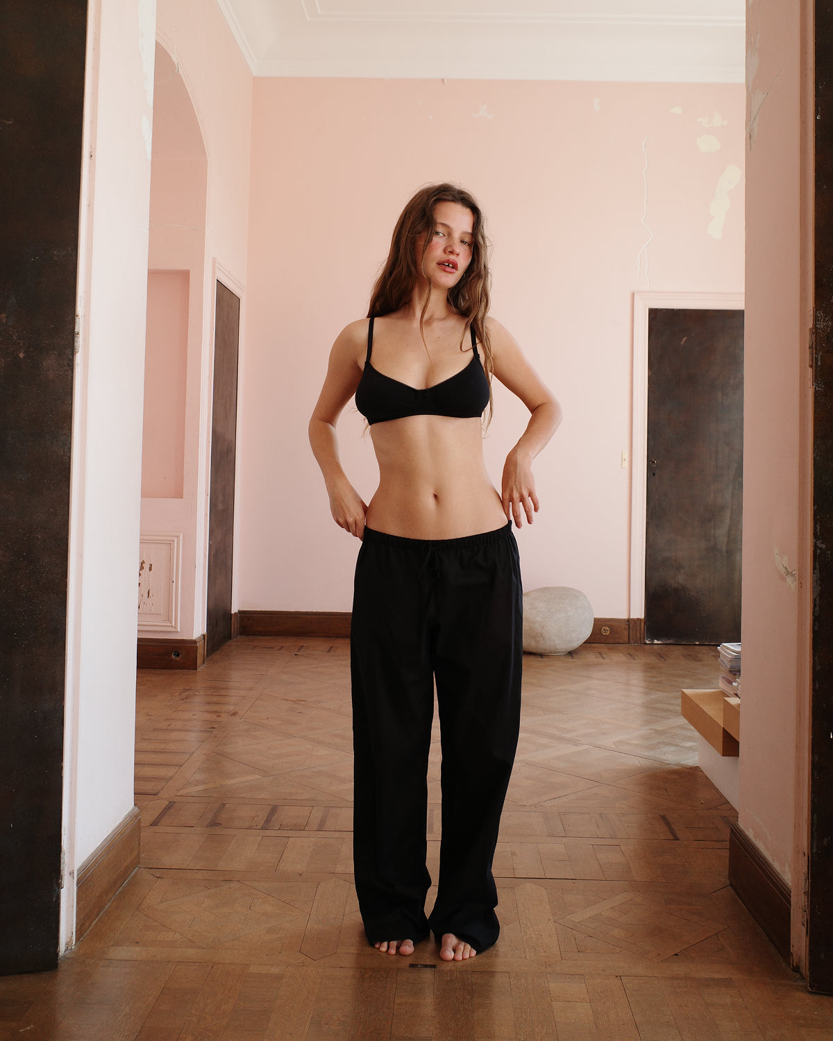 Woman wearing a black bra and black pants standing in a room with wooden floor and pink walls.