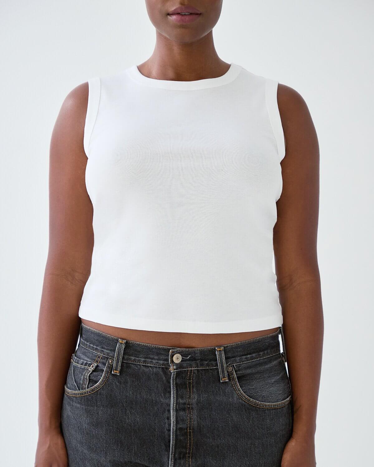 Person wearing a white sleeveless top and dark jeans on a plain background
